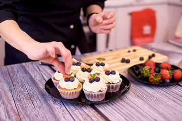 Cupcakes with strawberries and blueberries on a plate. Female hands fill cupcakes on the kitchen table.Selective focus.