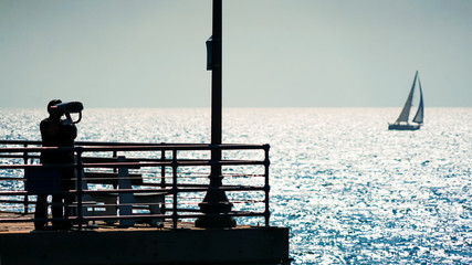 Mann beobachtet von einem Pier aus ein in der Ferne vorbeifahrendes Segelboot als Silhouette gegen...