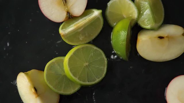 Fresh sliced fruits falling into water in slow Motion. Shot of organic limes and apples dropping in liquid with black background. Filmed in Prores.