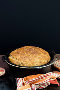 Sourdough Bread In Cast Iron With Colourful Tea Towel On Wooden Table With Black Background And Negative Space.