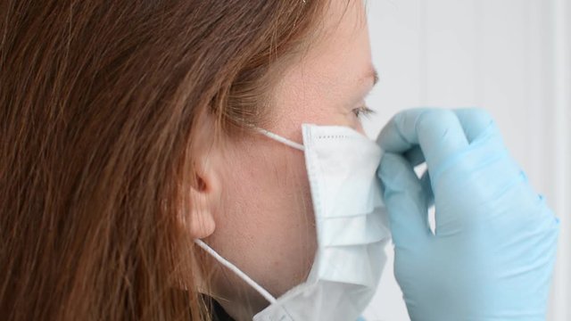 Red-haired girl puts on a disposable medical mask with hands in blue gloves, on a white background, close side view
