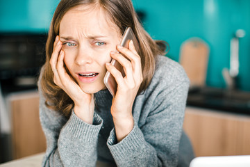 Closeup portrait of worried young beautiful woman talking on smartphone in kitchen. Bad news concept. Front view.