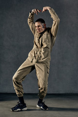 Active guy with tattooed body and face, earrings, beard. Dressed in khaki overalls and black sneakers. Dancing on gray background. Dancehall, hip-hop