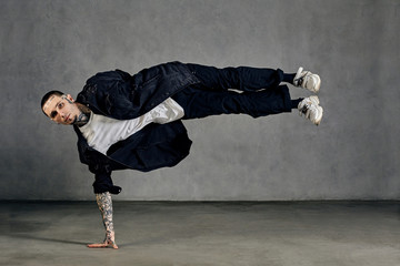 Fellow, tattooed body, beard. Dressed in white t-shirt and sneakers, black denim shirt, pants. Performing tricks, gray background. Dancehall, hip-hop