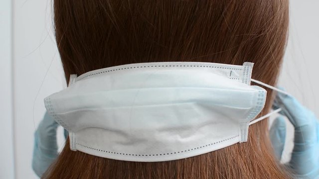 Medical mask shakes his head "no" on the back of the head of a red-haired long-haired girl held by hands in blue medical gloves on a white background in a static pose, close view