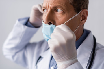 Fototapeta premium selective focus of middle aged doctor in latex gloves touching blue medical mask isolated on grey