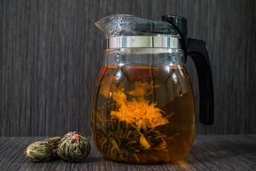 Blooming tea flower in glass teapot and dry flowering tea balls on brown wooden background.