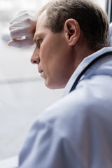 selective focus of tired middle aged doctor standing near window in clinic