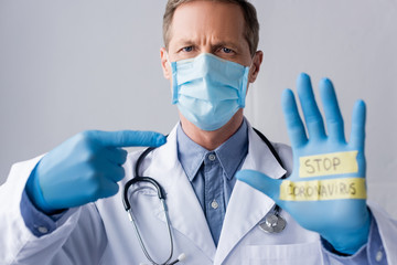 selective focus of mature doctor in medical mask pointing with finger at hand with stop coronavirus lettering isolated on grey