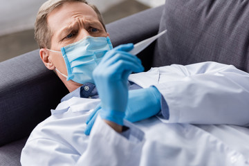 selective focus of mature doctor in medical mask and latex gloves looking at digital thermometer...