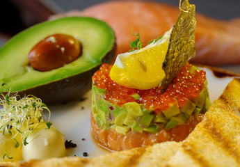 Tartar with red caviar,avocado and fish .The tartar stands on a white plate and next to it lies butter with fried bread.Decorated of fish scale and lemon.