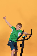 Joyful and happy child on a stationary bike raises his hands to the top and encourages sports and physical education.