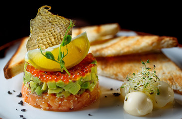 Tartar with red caviar,avocado and fish .The tartar stands on a white plate and next to it lies butter with fried bread.Decorated of fish scale and lemon.