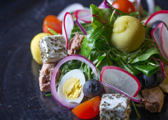 A salad of cherry tomatoes,arugula,lots of herbs,radishes,eggs,cheese and some meat.All the ingridients are on a dark table.