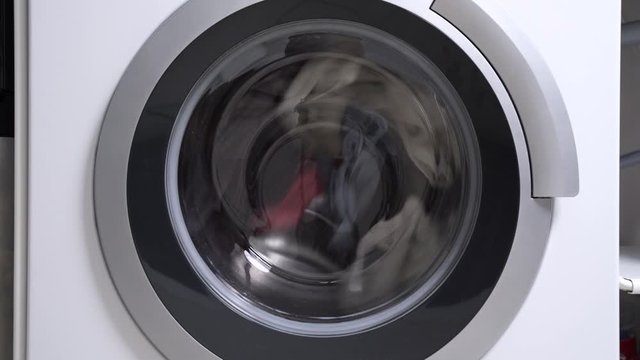Closed drum of the washing machine rotates slowly rinsing dirty multi-colored laundry with fabric softener, close up. Advertising of large household appliances for general cleaning of the house.
