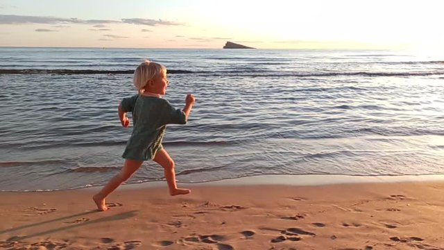 Blonde 3-years-old girl in knitted sweater laughing runs along water's edge on city's sandy beach at sunset. Long shot. Slow motion.