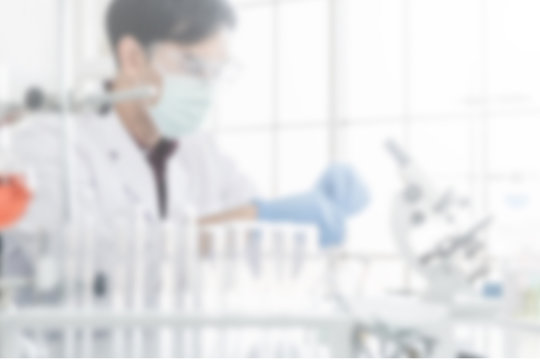 Blur out of focus photo of a male scientist with black hair wearing white coat protective glassware and face mask working with a microscope in a laboratory setting with test tubes.