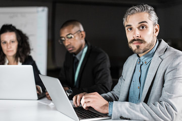 Concentrated manager sitting at table and looking at camera. Focused man working with laptop in office at night. Business, working late concept