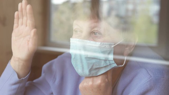 An elderly woman in a medical mask looks out the window. Quarantine self-isolation, coronavirus, covid 19.