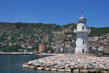 Lighthouse on the sea bank and fortress medieval tower far away. A view of the city on the mountain under clear blue skies.