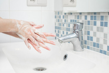 White woman washing her hand with foaming green soap in front of her white sink. her image is reflected in the mirror and the water flows through the silver tap. There is small tiling on the wall.