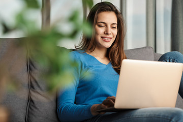 Image of happy woman using laptop and smiling while sitting on sofa