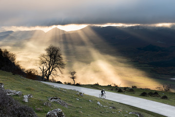 Fototapeta premium A romantic cyclist climbing a lonely road while the sunrise illuminates with beautiful light the valley, San Miguel de Aralar, Navarra, Spain.