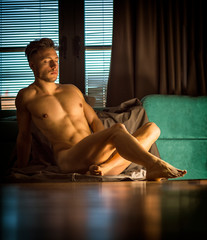 Handsome totally naked muscular young man sitting down on hardwood floor at home in seductive attitude, looking at camera