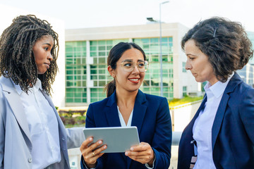 Positive female professional with tablet consulting colleagues and showing screen to them. Business women meeting and talking outside in city. Project discussion concept