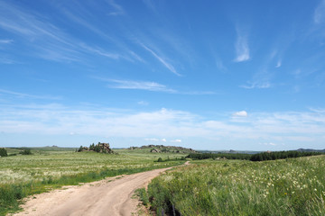 The landscape with the big ground road, the big field with green grass and flowers, the far rock, the white clouds in the blue sky.