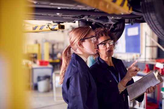 Female Tutor With Student Looking Underneath Car On Hydraulic Ramp On Auto Mechanic Course 