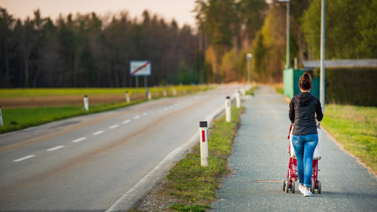 Young mother and child in the red stroller walks outdoor in rural area before sunset.