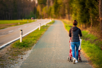 Young mother and child in the red stroller walks outdoor in rural area before sunset.