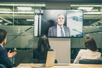 Obraz premium Employees listening to mature leader during video conference. Business people looking at monitor screen during video conference in office. Business conference concept