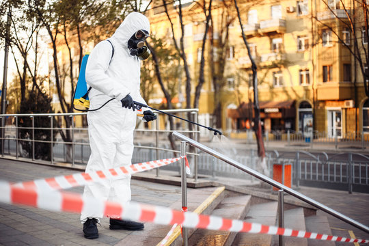 Man wearing personal protective equipment suit, gloves and gas mask cleaning the streets protected with warning tape with a backpack of spray disinfectant water to remove covid-19 coronavirus.