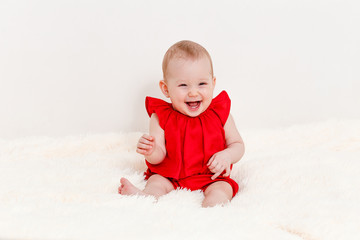 an eight-month-old cute Caucasian baby girl in a red suit sits on the bed and laughs. barefoot