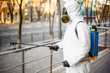 Man sprays disinfector onto the railing wearing coronavirus protective suit and equipment. Cleaning and sterilizing the not crowded city streets. Covid-19 nCov2019 spread prevention.