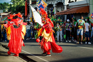 people in bright masquerade costumes walk by city street at dominican carnival