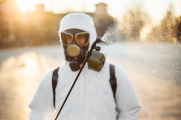 A man in a protective disinfection suit sprays sterilizer on the park square to prevent spread of coronavirus covid-19. Professional saniraty worker cleans city streets from the virus. Spot nCov2019.