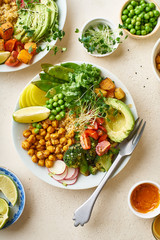 Healthy vegetarian lunch bowl with avocado, chickpeas, quinoa and vegetables, garnished with microgreens and nut dressing. Flat lay on stone background.
