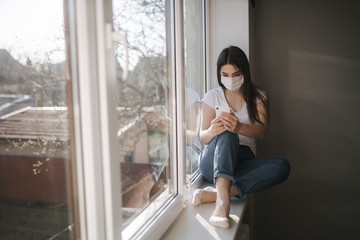 Fototapeta na wymiar Attractive woman in medical mask sits on windowsill at home. Female in white t'shirt and white medical mask use phone