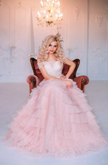 elegant beautiful young princess girl. Blonde queen woman sitting in old medieval vintage armchair. Golden crown pink vintage long dress. Backdrop shines crystal chandelier white interior classic room © kharchenkoirina