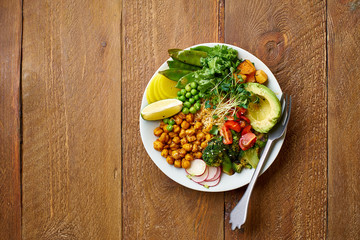 Healthy vegetarian lunch bowl with avocado, chickpeas, quinoa and vegetables, garnished with microgreens and nut dressing. Flat lay on wooden background.