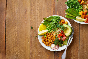 Healthy vegetarian lunch bowl with avocado, chickpeas, quinoa and vegetables, garnished with microgreens and nut dressing. Flat lay on wooden background.