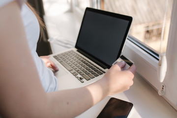 Close up of young woman shopping online using laptop phone and credit card. Laptop on windowsill
