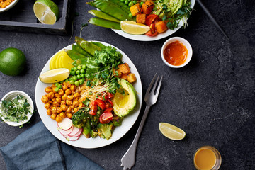 Healthy vegetarian lunch bowl with avocado, chickpeas, quinoa and vegetables, garnished with microgreens and nut dressing. Flat lay on dark concrete background.