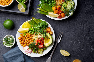 Healthy vegetarian lunch bowl with avocado, chickpeas, quinoa and vegetables, garnished with microgreens and nut dressing. Flat lay on dark concrete background.