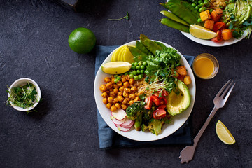 Healthy vegetarian lunch bowl with avocado, chickpeas, quinoa and vegetables, garnished with microgreens and nut dressing. Flat lay on dark concrete background.