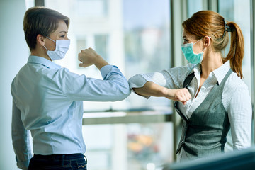 Female colleagues with face masks elbow bumping while greeting in the office. © Drazen