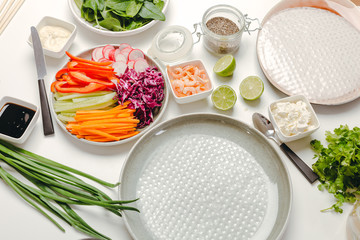 Side view of ingredients for cooking spring rolls - carrots, cucumbers, herbs, peppers, red cabbage, shrimps, rice paper and soy sauce on a white background.